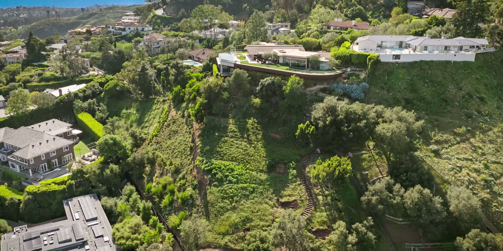 Aerial view of a green hillside parcel above the Pacific Palisades with neighboring homes on the ridge